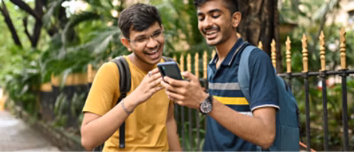 Two young men laughing and looking at a smartphone together, standing outdoors with greenery in the background.