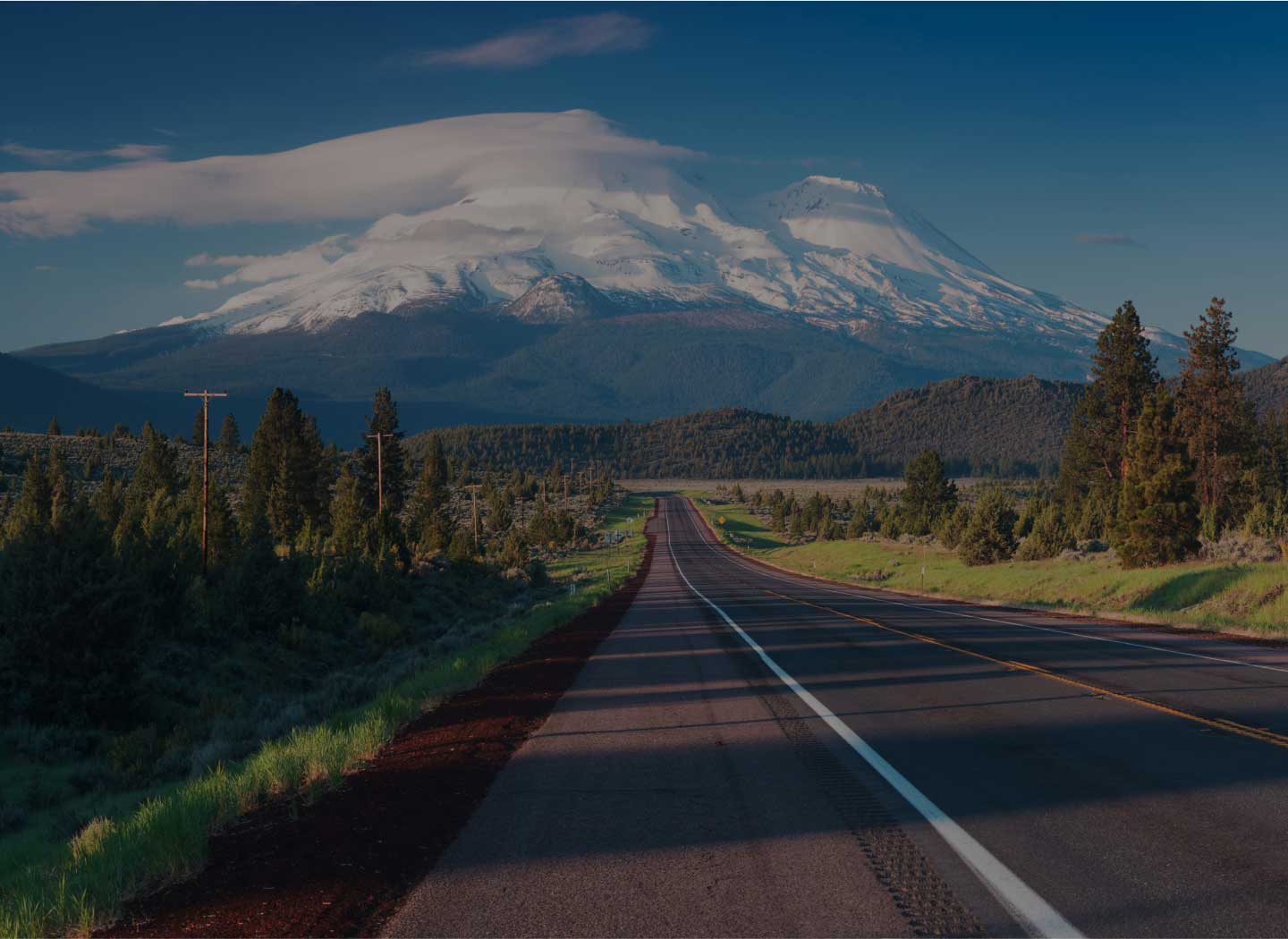 Scenic highway leading toward a snow-capped mountain, surrounded by lush greenery and tall trees.